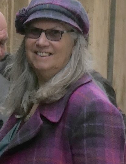 women with mid long grey hair wearing a purple cap and coat, smiling. Wearing sunglasses